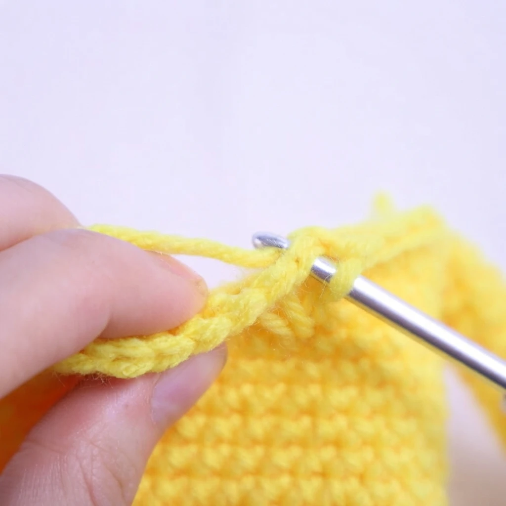 Hands holding yellow crocheted fabric while a metal crochet hook is inserted into a stitch, showing a step of a tight single crochet technique