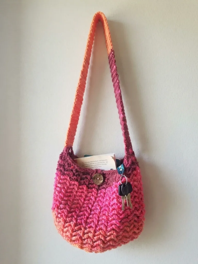 Crocheted pink and orange striped shoulder bag with button closure holding keys and a card, hanging on a wall.