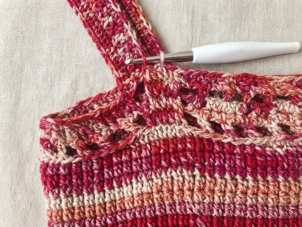 Close-up of a striped crochet market bag in progress, with red, pink, peach and cream yarn, openwork mesh, a strap, and a white-handled crochet hook in the work.