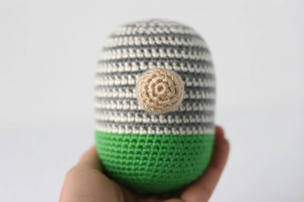 Crochet gnome body held in a hand, green at the bottom with grey and white stripes above and a tan round nose in the center