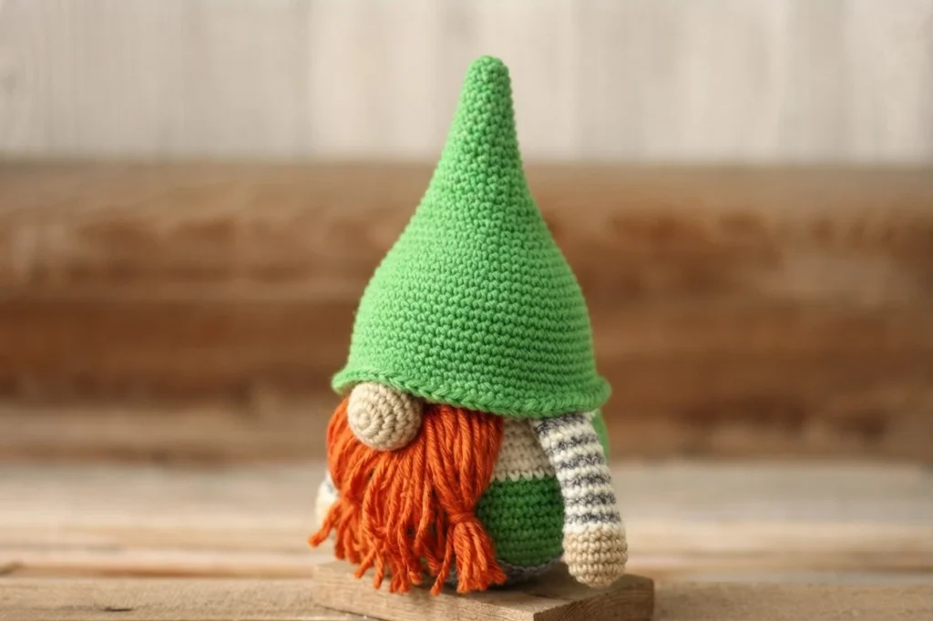 Crochet gnome in three-quarter view with a tall green pointed hat, beige round nose, long orange beard and a grey and white striped arm, standing on a wooden plank
