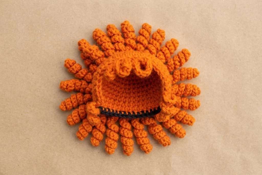 Orange crocheted hair cap with short bangs at the front and many curled strands around the edge, laid flat on a plain tan surface