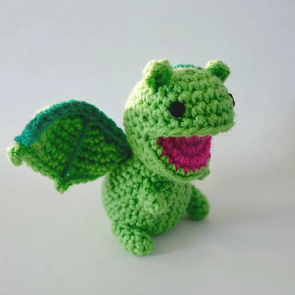 Green crocheted dragon amigurumi with open pink mouth, small ears and one teal rimmed wing spread, shown on a white background.