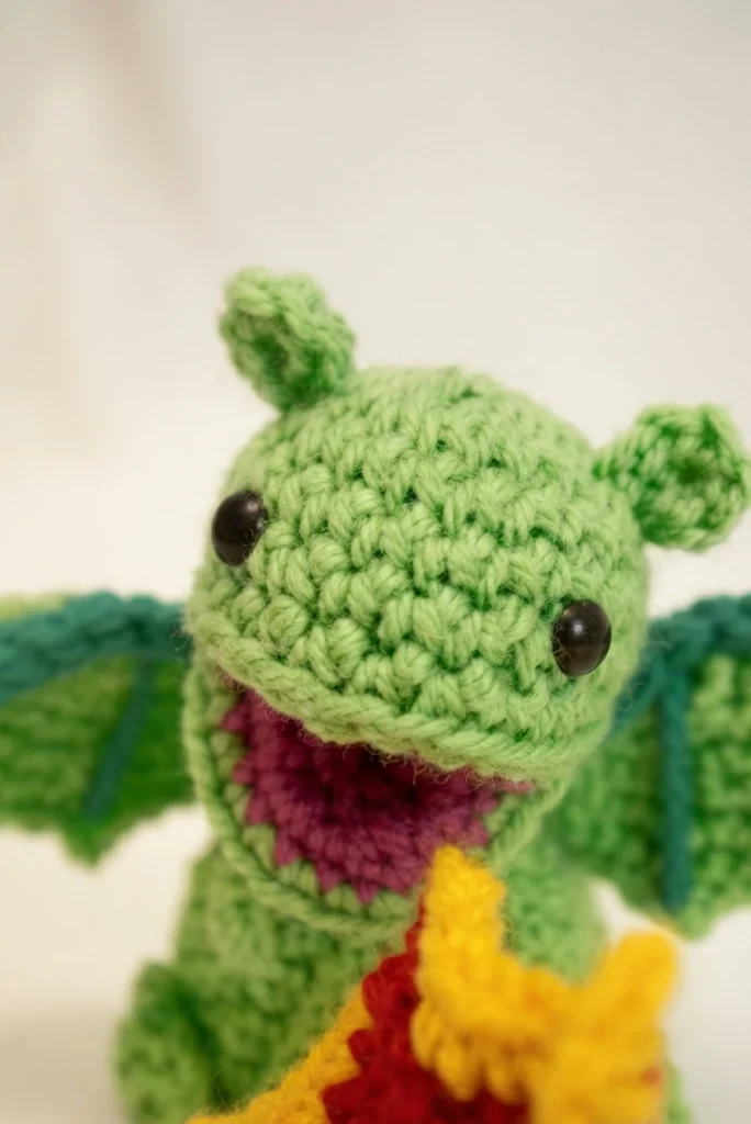 Close-up of a crocheted green dragon amigurumi with an open pink mouth, round black eyes and teal wing edges, a red and yellow crocheted flame at its mouth.