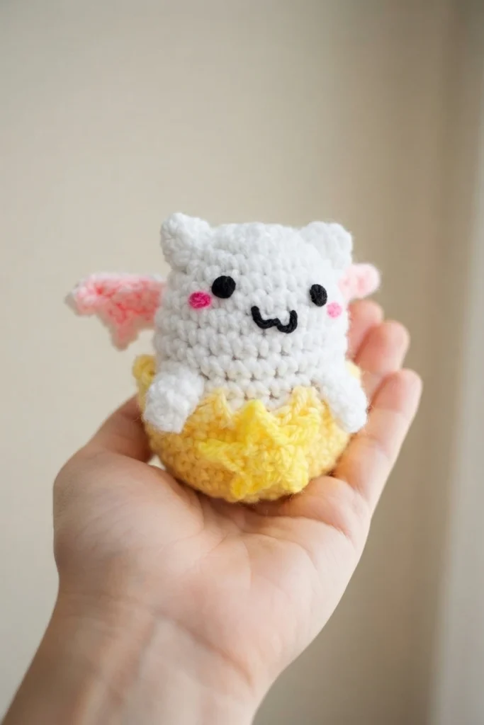 Hand holding a small white crocheted amigurumi with pink wings and embroidered face, sitting inside a cracked yellow crocheted egg