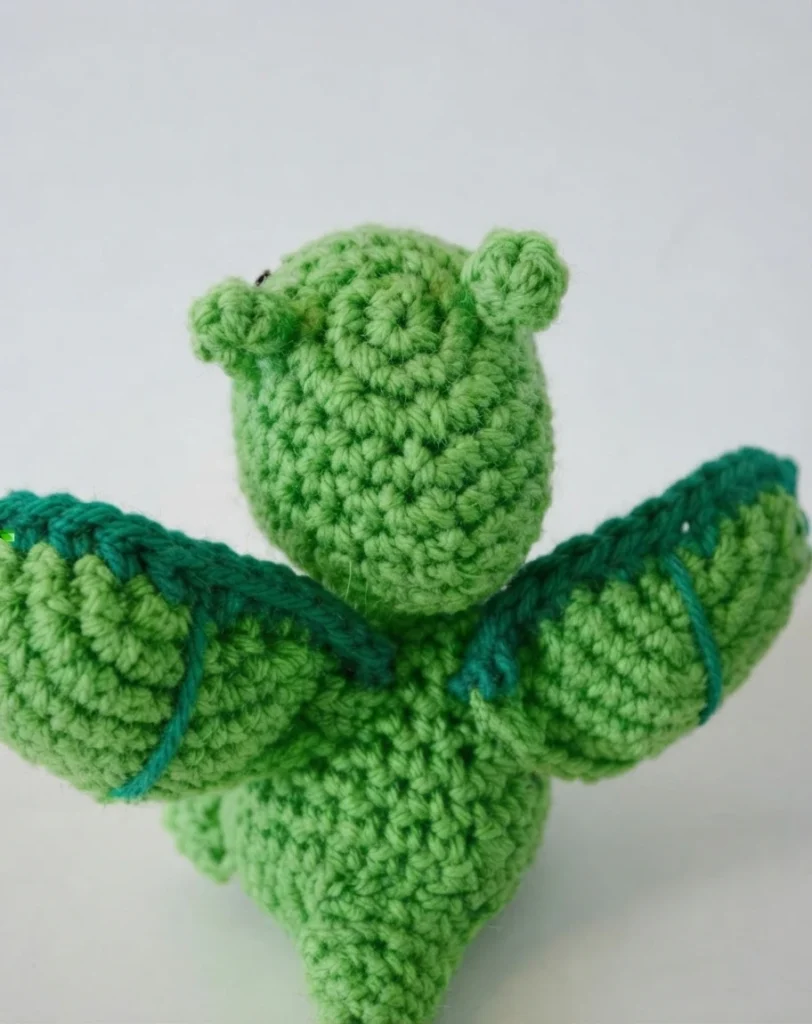Back view of a small green crochet dragon amigurumi with wings spread, edged with a darker teal rim