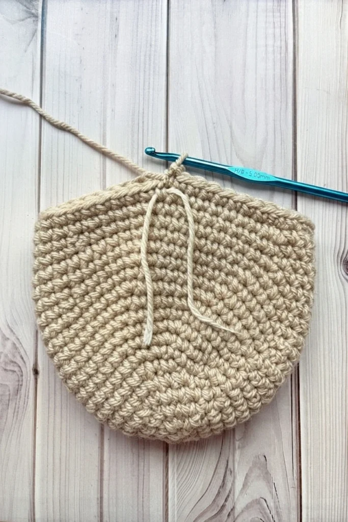 A beige crocheted piece with a loose yarn strand and a blue crochet hook on a pale wood surface.