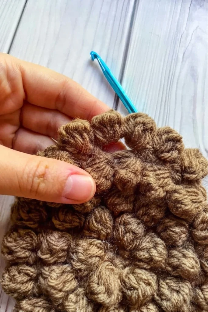 A hand holds open the brown bobble-textured crochet top around a small gap, with a blue crochet hook above it.