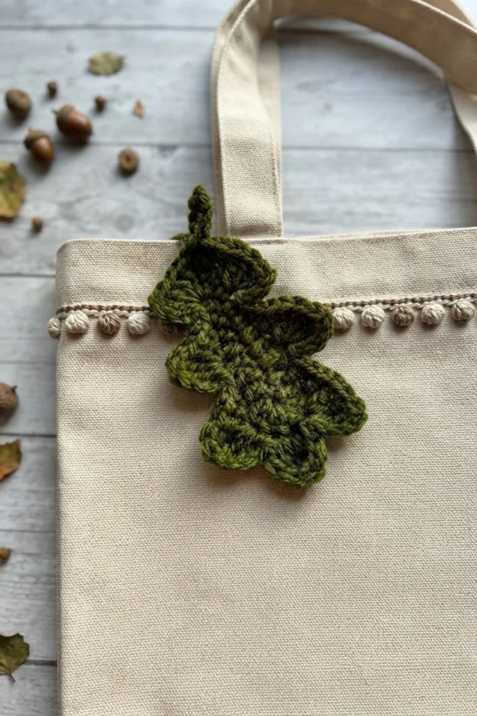 Feuille de chêne crochetée en vert, fixée sur un sac en toile crème orné d’une bordure à pompons.