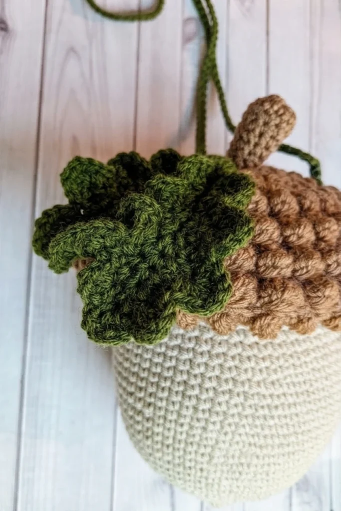 A cream crocheted acorn bag with a textured brown top, a prominent green leaf and a long strap against a pale wooden background.