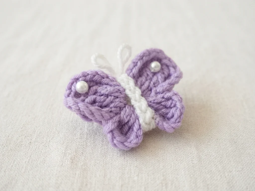 A small crochet butterfly with purple wings, a white body, looped white antennae and a pearl bead on each upper wing, resting on white fabric.
