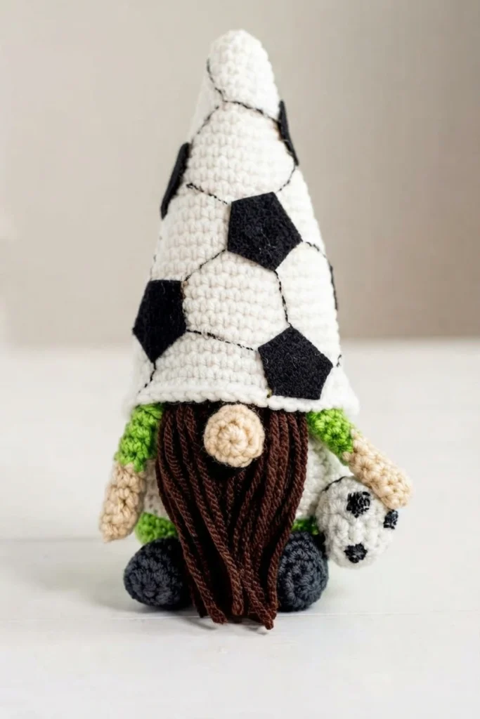 A crochet soccer gnome with a tall white cone hat patterned like a soccer ball with black pentagons, a round skin-tone nose, a long dark brown beard, green shoulders, small hands and dark feet, holding a tiny crocheted soccer ball.