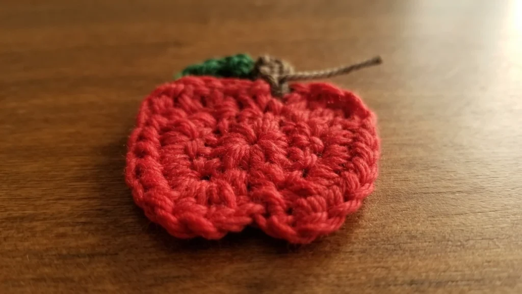 Crocheted red apple applique with a brown stem and a small green leaf, lying flat on a wooden surface