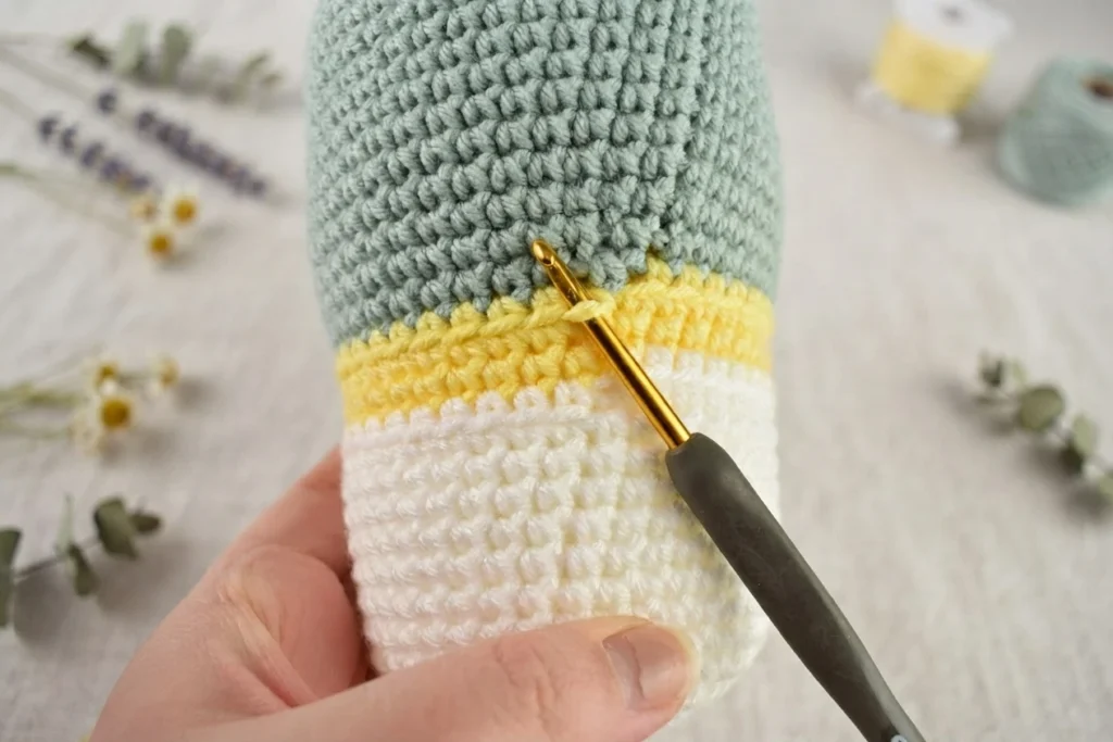 Close-up of a hand holding a crocheted gnome hat in progress, showing white, yellow and sage green striped rounds with a gold crochet hook inserted at the color join.