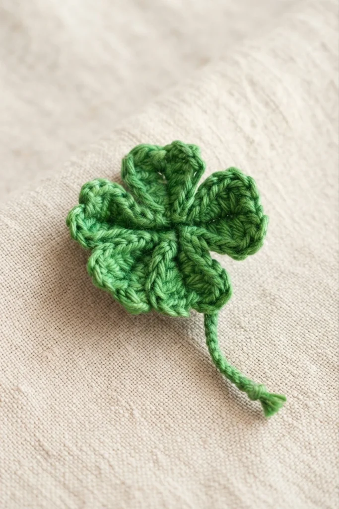 Green crocheted four-leaf clover with a twisted yarn stem resting on beige linen fabric.