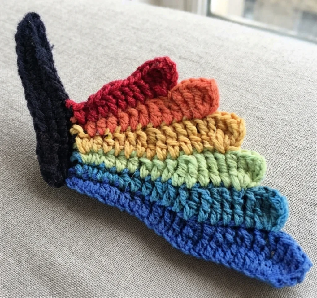 Partially crocheted butterfly wing with layered rainbow stripes joined to a black body section, resting on beige fabric.