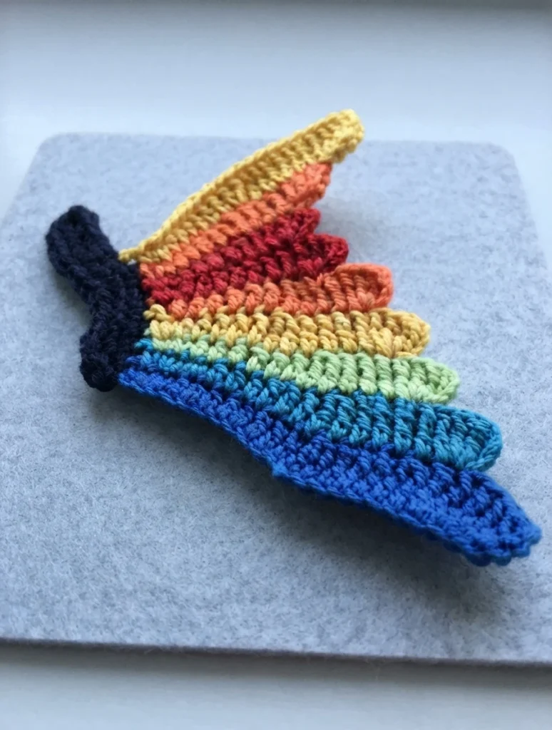 Crocheted butterfly wing with nine rainbow-colored segments joined to a black body piece, photographed on gray felt
