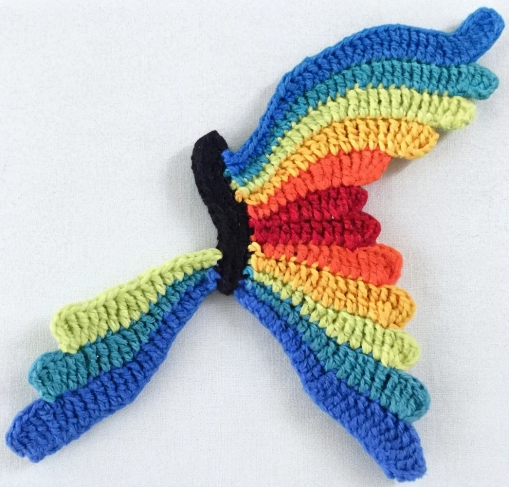 Rainbow-striped crocheted butterfly wing partially worked and joined to a black crocheted body, shown on a light plain background.