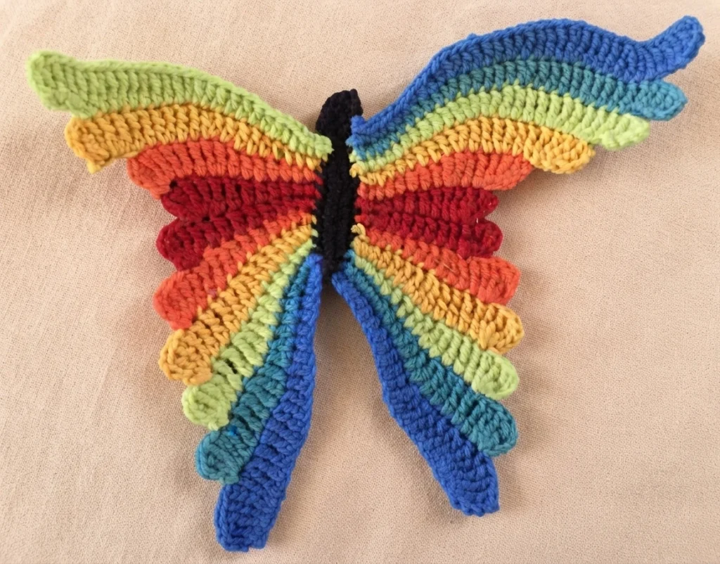 Rainbow-striped crochet butterfly with black body lying flat on a beige fabric background.