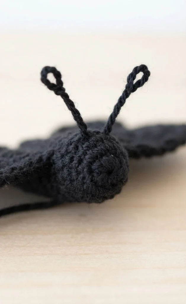 Close-up of a black crocheted butterfly head with two looped antennae, a dark wing softly blurred behind it on a light wooden surface.