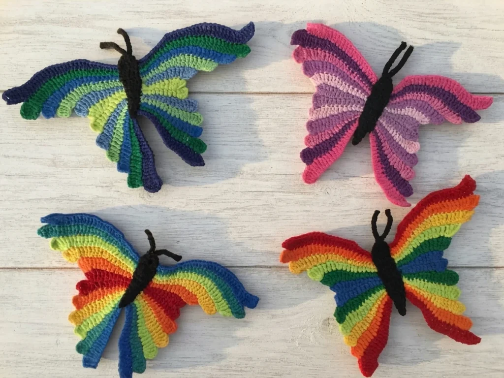 Four crochet butterflies in blue-green, pink-purple and two rainbow colorways arranged on a whitewashed wooden surface