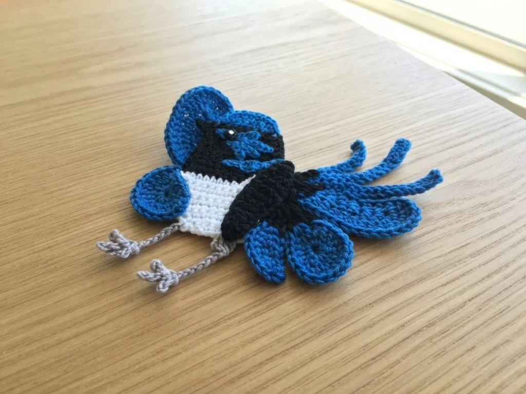 Flat crochet blue wren applique with blue head markings, black chest, white belly and grey legs, lying sideways on a light wooden table