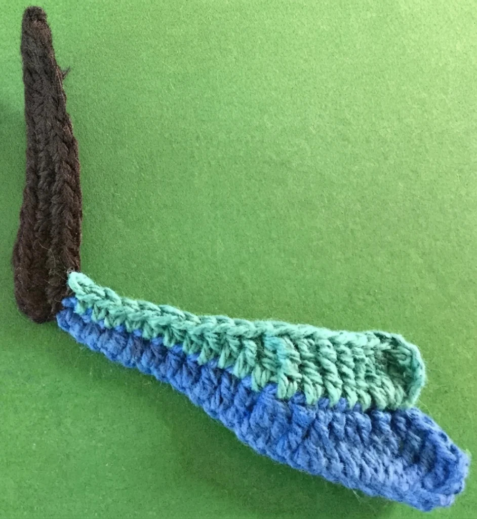 Partial crochet butterfly wing in progress with a dark brown body and two wing segments in royal blue and teal green, lying on a green background.