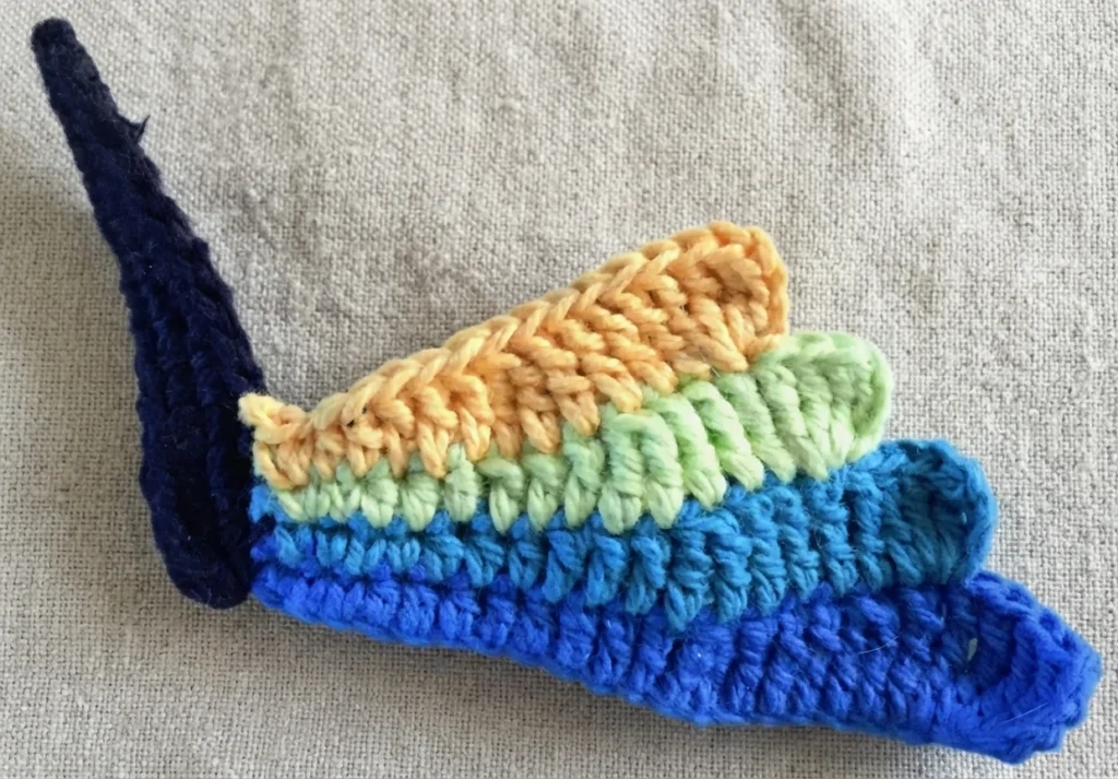 Close-up of a crochet butterfly wing in progress: dark navy body section with four overlapping wing segments in blue, teal, green and pale orange on a beige fabric background