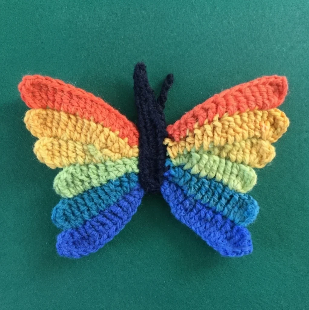 Crocheted butterfly with rainbow striped wings and a dark body on a green background.