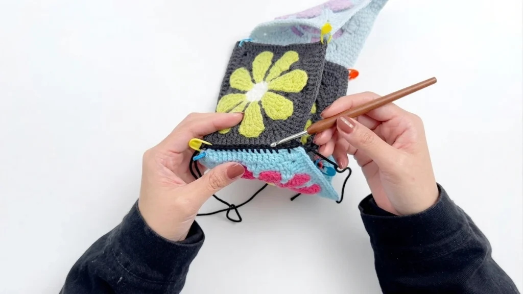Hands seaming two flower granny squares together with black yarn and a wooden crochet hook, stitch markers along the edges, on a white background