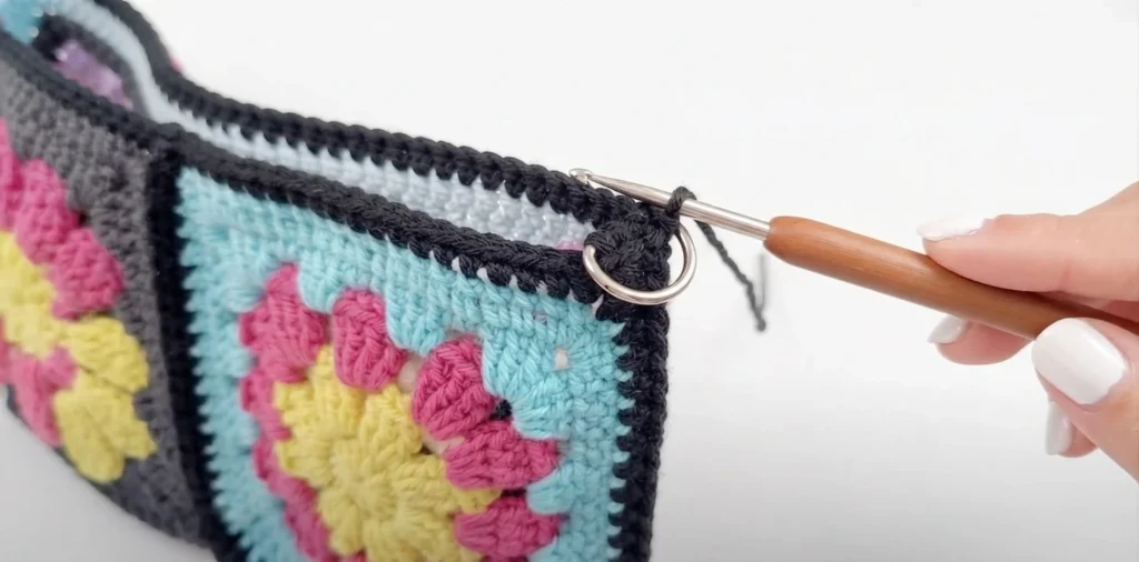 Close-up of a hand crocheting black edging around the opening of a colorful granny square bag with a metal D-ring at the corner
