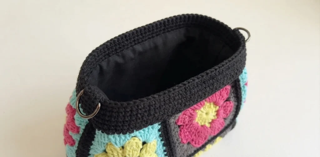 Open crochet granny square bag showing the finished black single crochet top edge with metal D-rings, pink and yellow flower motifs on the sides