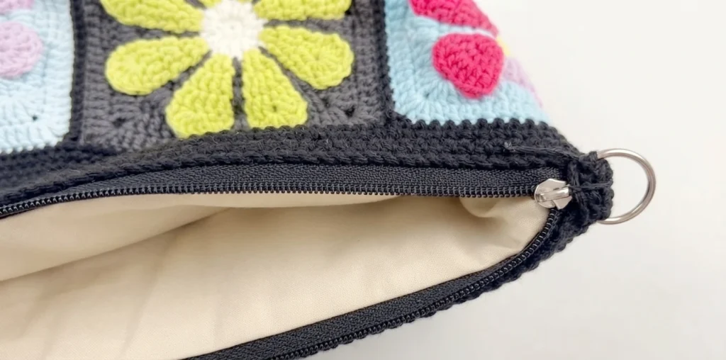 Close-up of a crochet granny square crossbody bag with floral motifs in green, pink and purple on black and blue squares, zipper open revealing a beige fabric lining.