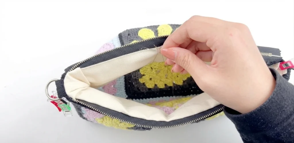 Hand stitching the cream fabric lining to the zipper inside a crochet granny square crossbody bag