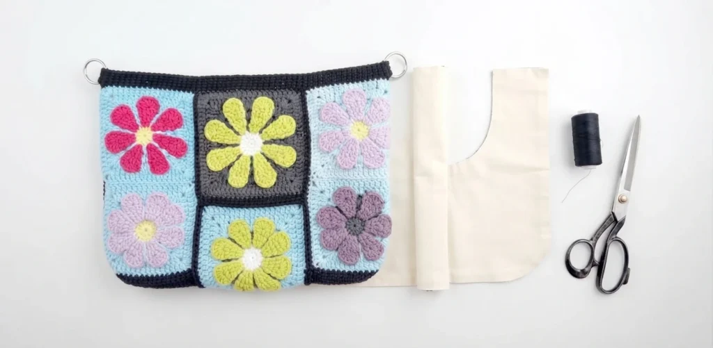 Flat lay of a granny square crochet bag panel with colorful flower motifs beside a cut lining fabric, a thread spool and scissors.
