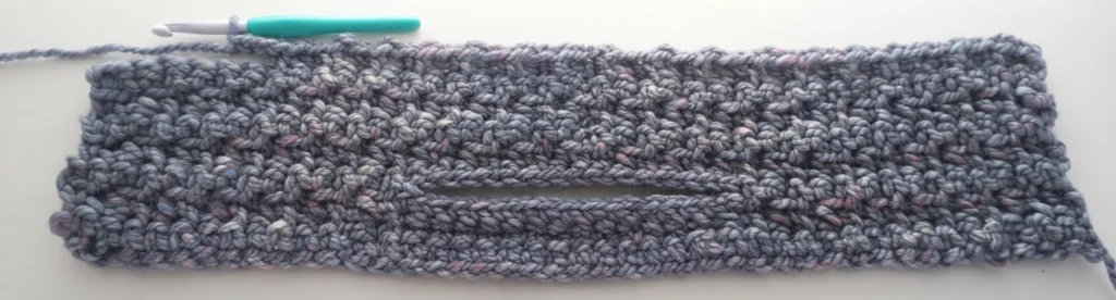 Crocheted front panel in chunky grey-blue yarn with pink flecks, lying flat on a white surface, showing a horizontal opening for a hidden pocket, a teal crochet hook resting near the top edge and a working loop of yarn at the left.