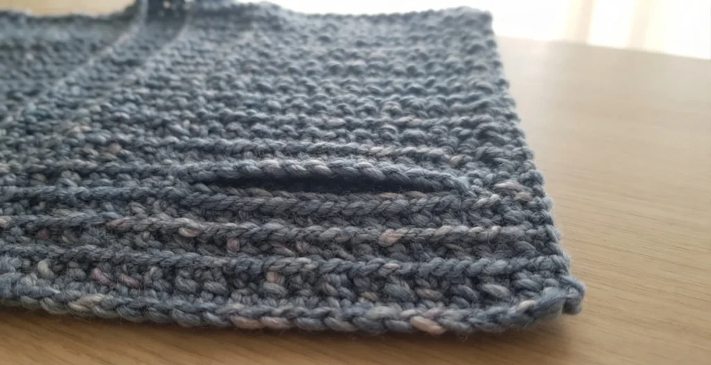 Close-up of a chunky gray crochet panel with a horizontal slit opening for a pocket, resting flat on a light wood table.