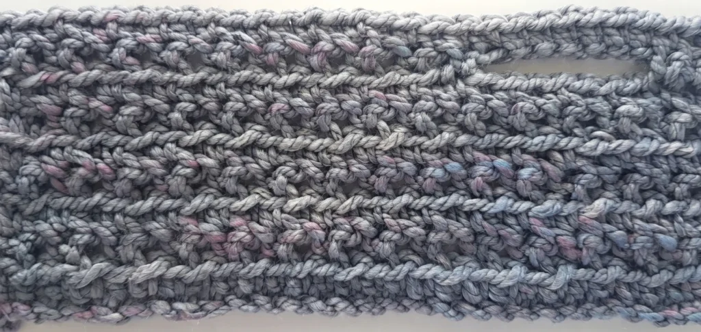 Macro view of a chunky grey-blue crochet panel worked in an open mesh stitch, with a horizontal slit pocket opening near the top right and pink and purple flecks visible in the yarn.
