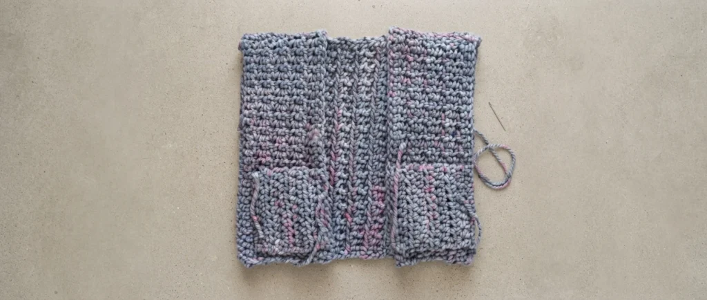 Flat lay of a chunky crochet cardigan back panel with two front panels laid over it, both fronts showing small pocket openings near the lower edge, with a tapestry needle and a loop of yarn resting beside the pieces on a concrete surface.