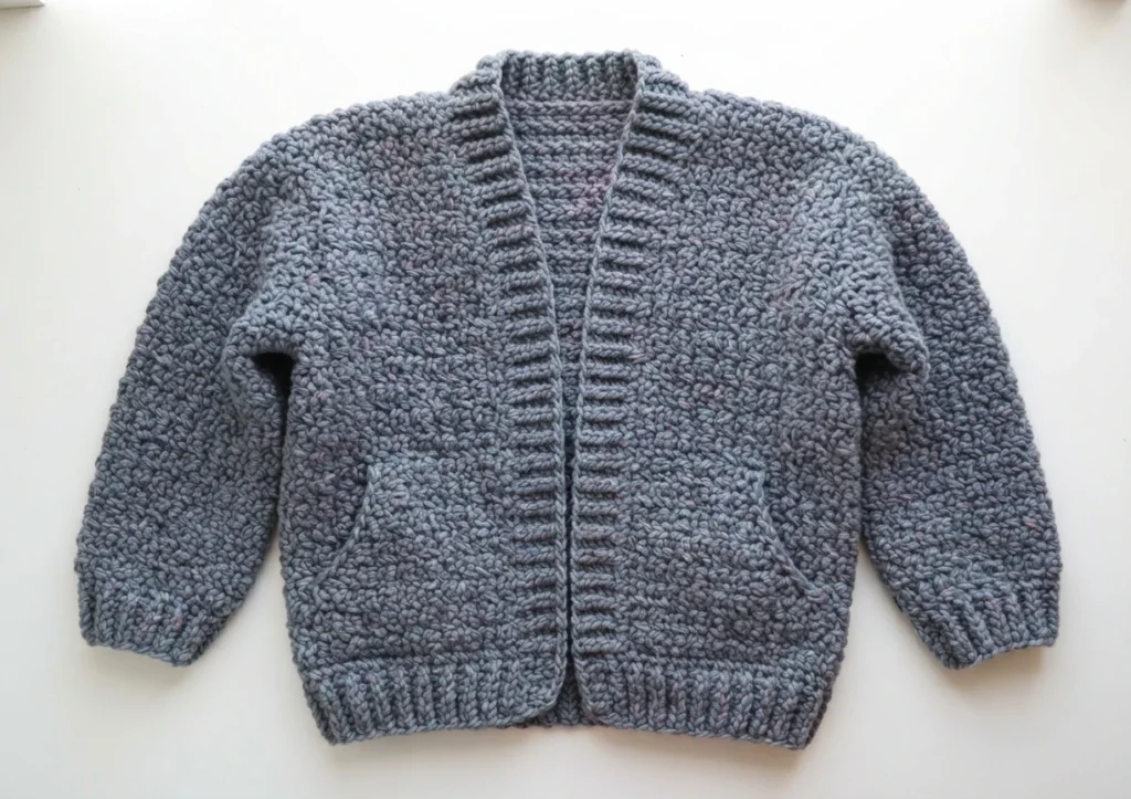 Chunky grey-blue marled crochet bomber cardigan laid flat, showing ribbed neckline and front bands, ribbed cuffs and hem, two front pockets and textured mesh body.