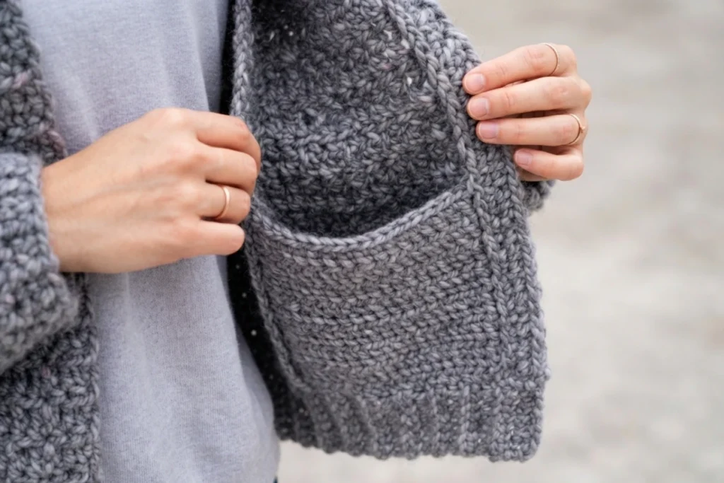 Close-up of two hands holding open the hidden pocket of a chunky gray crochet cardigan worn over a gray top