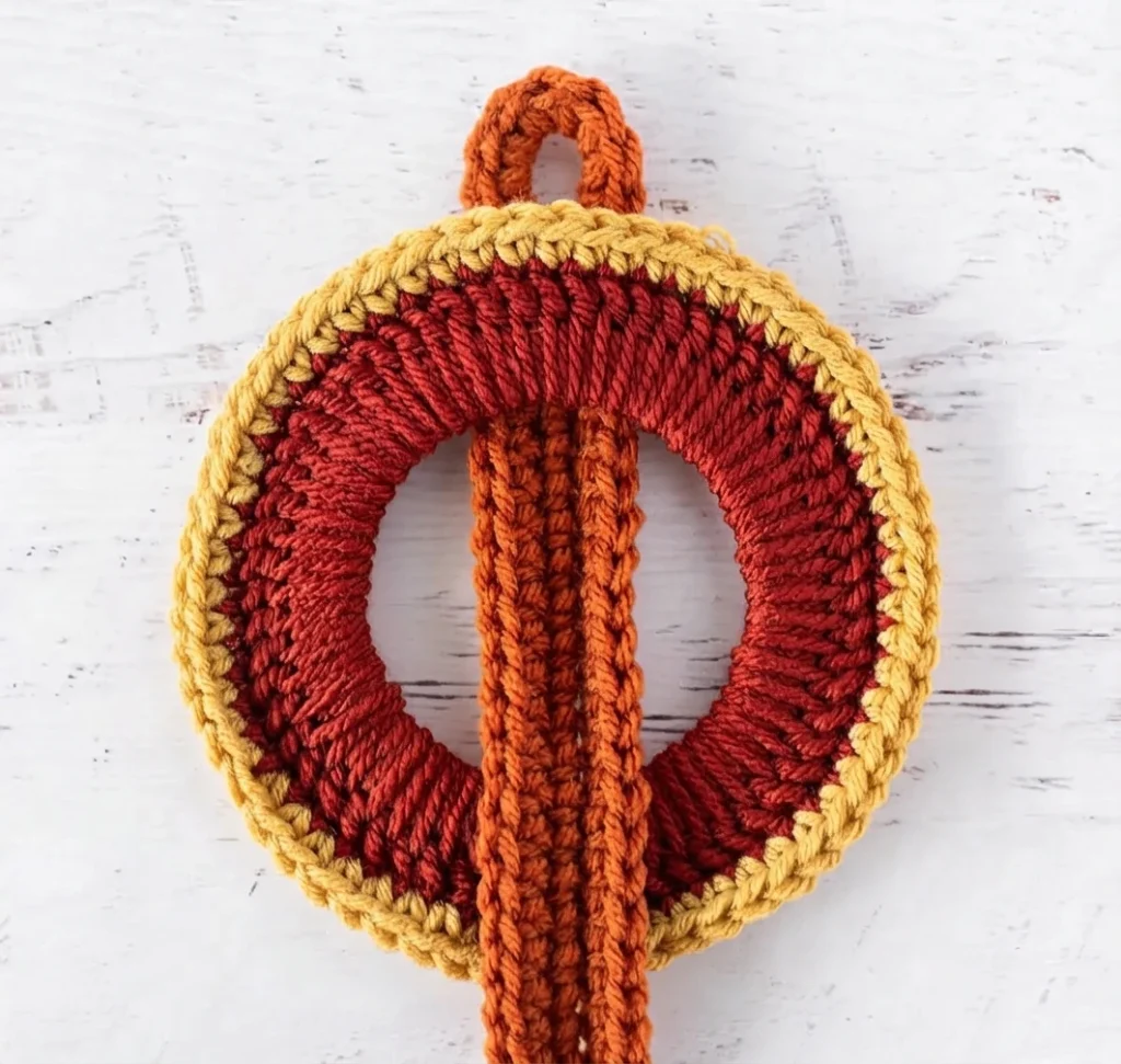 Gros plan d’un anneau crocheté rouge foncé bordé de jaune, traversé verticalement par une sangle orange et surmonté d’une boucle.