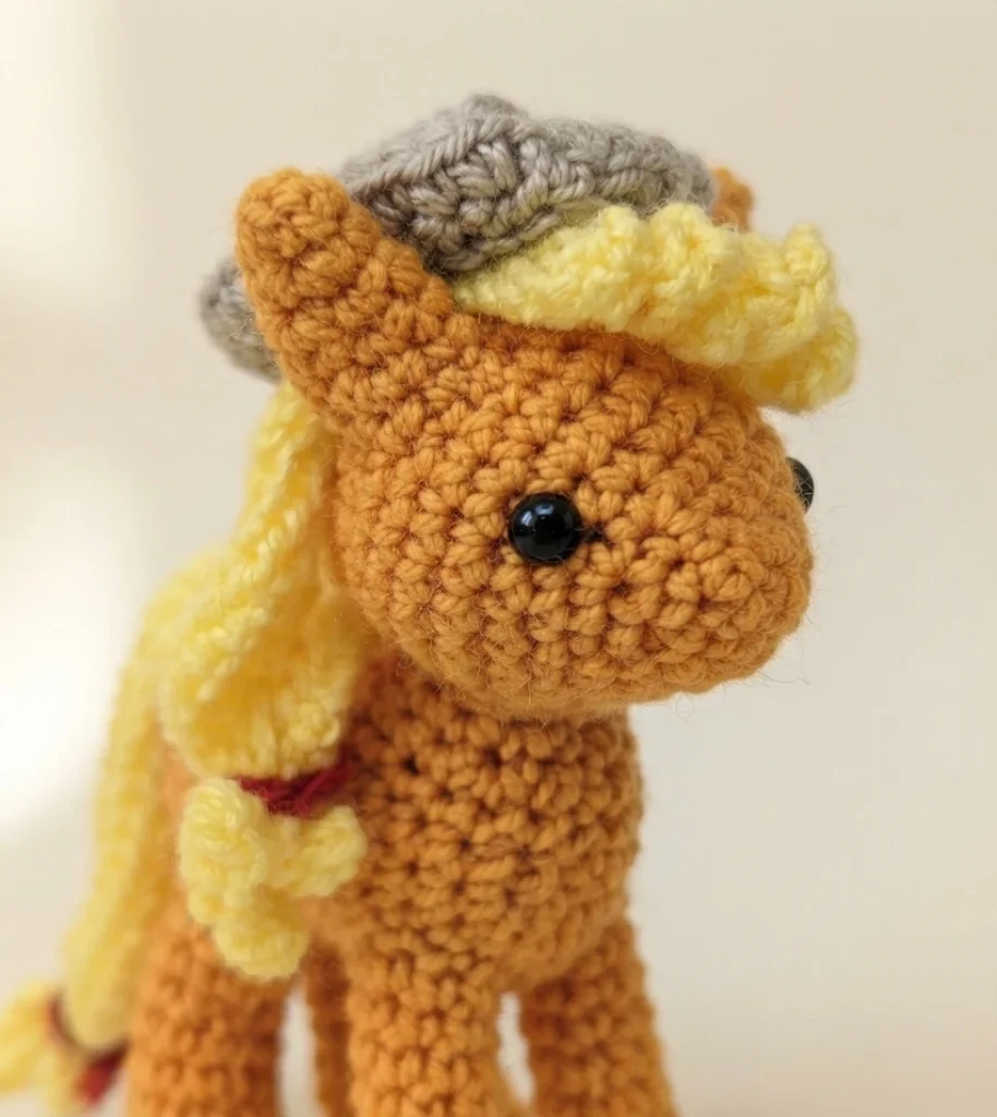 Close-up of a finished orange crocheted amigurumi pony head with yellow mane, gray crocheted hat and black plastic safety eyes