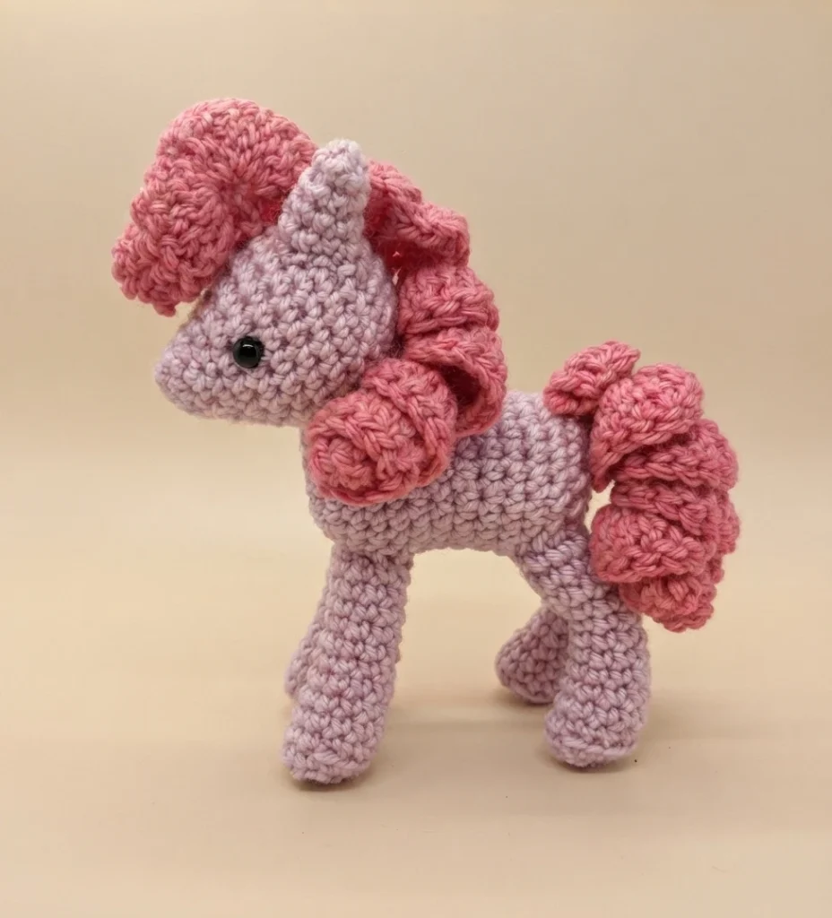 A crocheted amigurumi pony with a pale lavender body, curly pink mane and tail, and black safety eyes, shown standing in profile on a plain background.