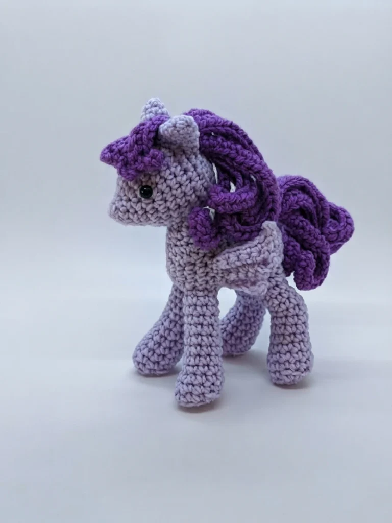 A finished lavender crocheted pony amigurumi with a curly dark purple mane and tail and black safety eyes, standing on a white background.
