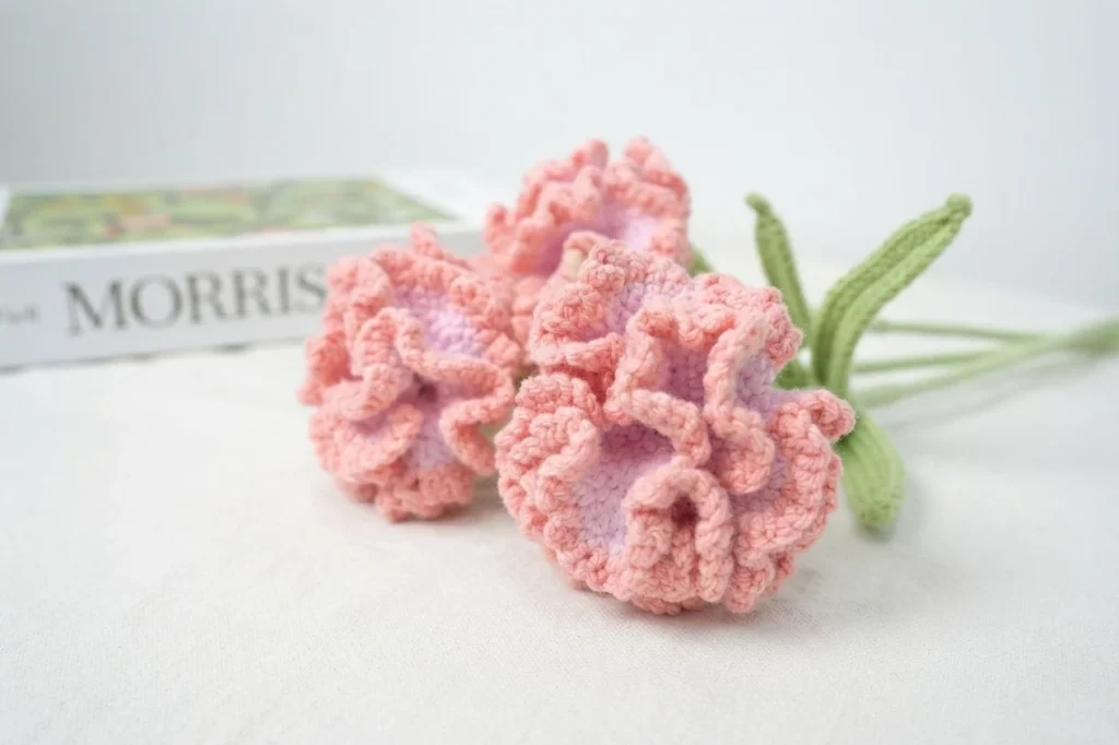 Three pink crochet carnations with ruffled petals and green stems lying on a white surface with a book in the background