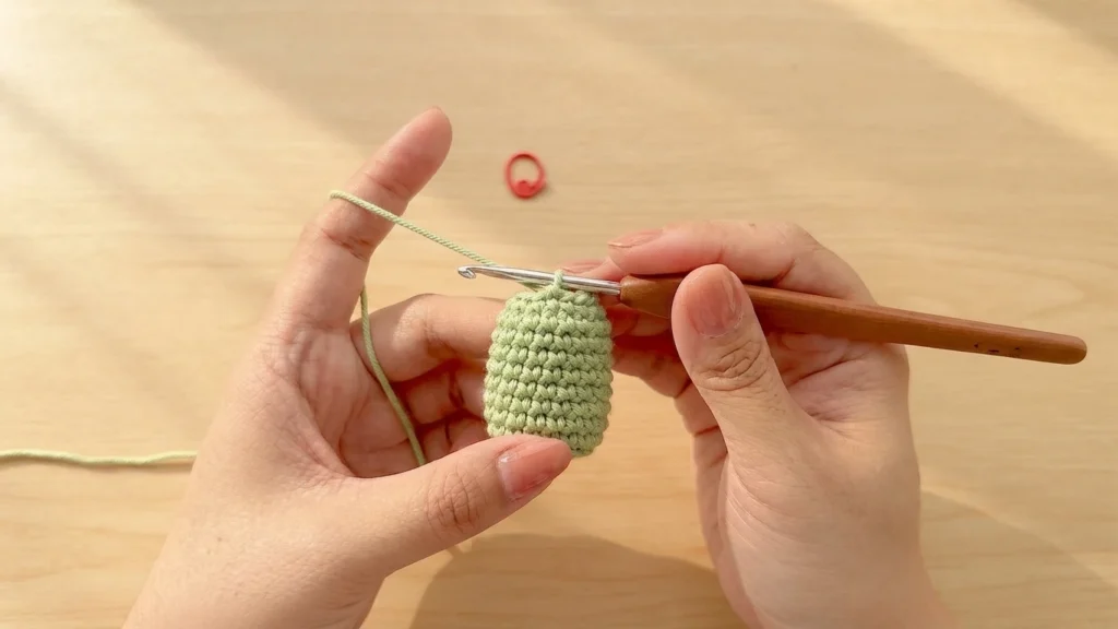Hands crocheting a small light green cylindrical flower base with a wooden handled hook, with a red stitch marker nearby