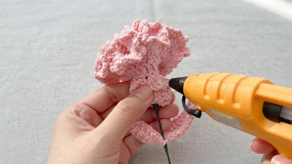 A hand holding a pink ruffled crochet carnation while a yellow hot glue gun applies glue to the folded base on a green wire stem