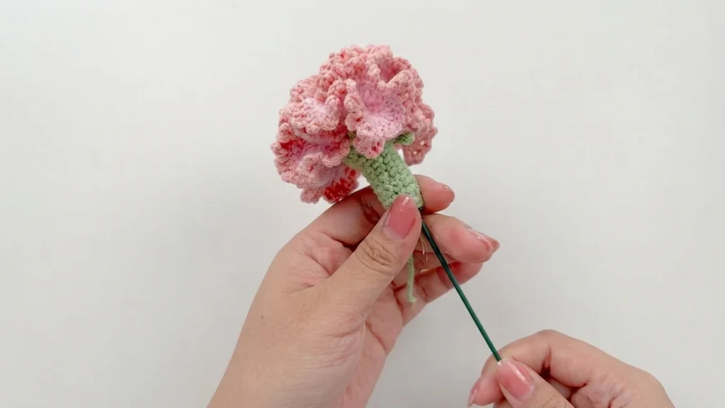 Hands inserting a dark green wire stem into the green base of a pink ruffled crochet carnation