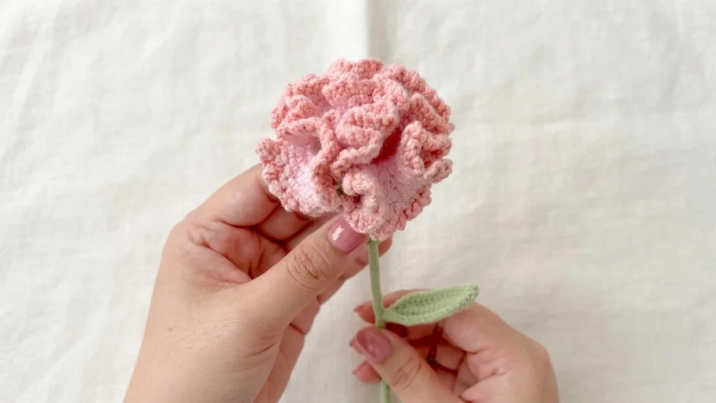 Hands holding a single pink ruffled crochet carnation with a green stem and leaf against a light fabric background